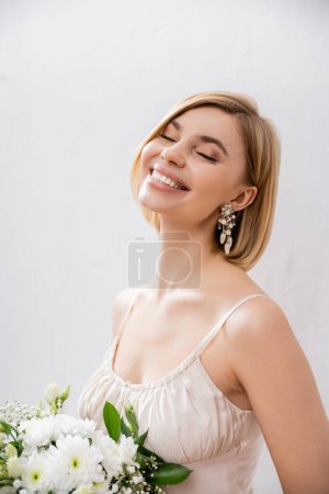 schöne und blonde Braut im Brautkleid mit Blumenstrauß auf grauem Hintergrund, Freude, weiße Blumen, Brautzubehör, Glück, besonderer Anlass, feminin, selig 