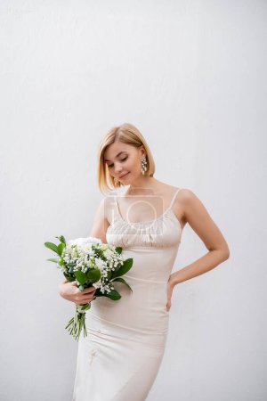glückliche und blonde Braut im Brautkleid mit Blumenstrauß auf grauem Hintergrund, weiße Blumen, Brautzubehör, Glück, besonderer Anlass, schön, feminin, selig 