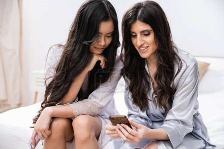 wedding preparations, happy interracial women in silk robes, using smartphone, digital age, bride and bridesmaid during slumber party, pretty girls, best friends, cultural diversity  