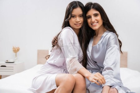 bridal party, bride with her bridesmaid sitting on bed, hugging each other, silk robes, best friends, brunette hair, cultural diversity, multiracial women, wedding planning, looking at camera