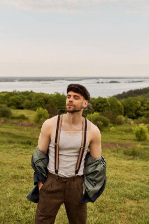 Fashionable and bearded man in jacket, suspenders and newsboy cap holding hands in pockets and looking away while standing with blurred nature at background, fashion-forward in countryside