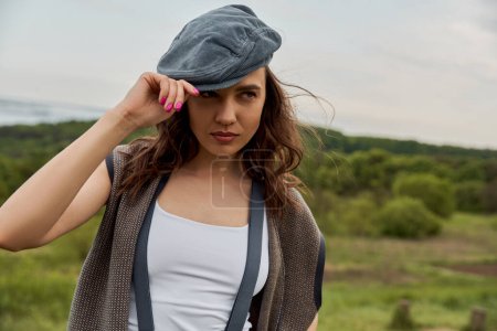 Fashionable brunette woman in suspenders and vintage clothes wearing newsboy cap and looking away while standing with blurred nature at background, fashion-forward in countryside