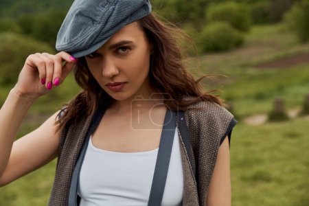 Portrait of stylish brunette woman in vintage clothes, vest and suspenders touching newsboy cap and looking at camera with blurred landscape at background, fashion-forward in countryside