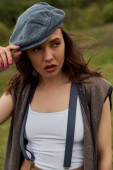 Portrait of fashionable brunette woman in vest and vintage suspenders touching newsboy cap and looking away while standing at blurred nature, fashion-forward in countryside puzzle #663007964