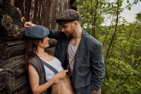 Cheerful and fashionable woman in newsboy cap touching suspender on bearded boyfriend in jacket while standing near rustic house with nature at background, stylish couple in rural setting