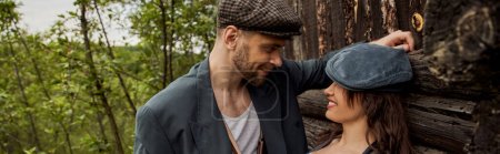 Smiling and fashionable man in jacket and newsboy cap looking at cheerful girlfriend while standing together near rustic house at nature, stylish couple in rural setting, banner 
