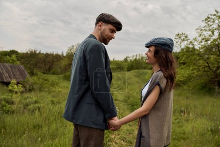 Bearded and stylish man in jacket and newsboy cap holding hand and looking at cheerful girlfriend in vest and standing together on grassy lawn at overcast, stylish couple in rural setting
