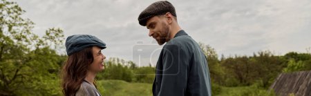 Photo for Cheerful brunette and stylish woman in newsboy cap looking at bearded boyfriend in jacket while standing together with landscape and overcast at background, stylish couple in rural setting, banner - Royalty Free Image