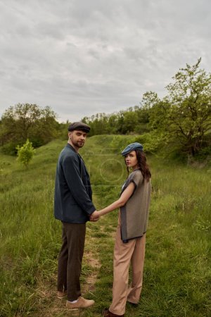 Fashionable bearded man in jacket and newsboy cap holding hand of brunette girlfriend and looking at camera with landscape and overcast at background, stylish couple in rural setting