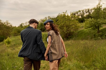 Fashionable brunette woman in vest and newsboy cap looking at camera and holding hand of bearded boyfriend and walking with landscape at background, stylish couple in rural setting