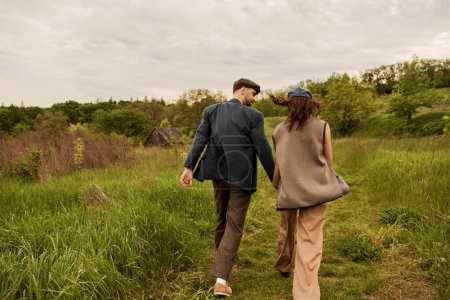 Bearded and stylish man in jacket and newsboy cap holding hand and looking at brunette girlfriend in vest while walking on meadow with nature at background, stylish couple in rural setting