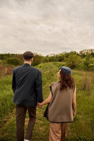 Brunette et femme à la mode en gilet et bonnet de newsboy tenant la main du petit ami en veste tout en marchant ensemble sur la prairie avec la nature et le ciel en arrière-plan, avant-garde de la mode dans la campagne