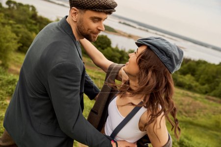 Cheerful and fashionable brunette woman in vintage suspenders and vest hugging bearded boyfriend in jacket and newsboy cap with landscape at background, fashion-forwards in countryside