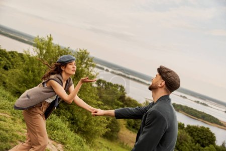 Femme brune à la mode en bonnet de newsboy et gilet soufflant baiser d'air et tenant la main du petit ami barbu en veste avec la nature et le ciel en arrière-plan, avant-garde de la mode dans la campagne