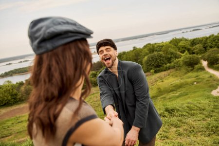 Cheerful and trendy bearded man in jacket and newsboy cap holding hand of blurred girlfriend while standing with scenic landscape and sky at background, fashion-forwards in countryside