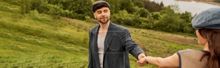 Uśmiechnięty i modny brodaty mężczyzna w czapce i kurtce z gazeciarzem trzymający za rękę zamazaną brunetkę podczas spędzania czasu z naturą w tle, modna para na wsi, baner 