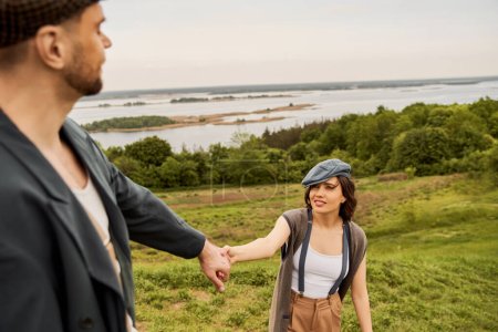 Photo for Smiling and trendy brunette woman in suspenders and newsboy cap holding hand of blurred boyfriend in jacket while standing with nature and sky at background, fashionable couple in countryside - Royalty Free Image