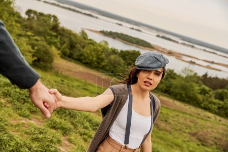 Fashionable brunette woman in newsboy cap and suspenders holding hand of blurred boyfriend and looking at camera with scenic landscape at background, fashionable couple in countryside
