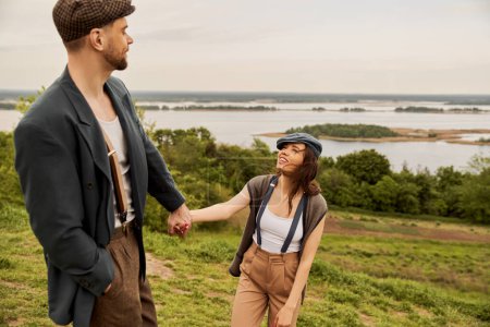 Cheerful and fashionable brunette woman in newsboy cap and suspenders holding hand of bearded boyfriend in jacket and standing with scenic landscape at background, fashionable couple in countryside