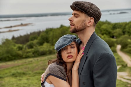 Brunette and fashionable woman in newsboy cap and vest hugging bearded boyfriend in jacket and standing with scenic landscape at background, fashionable couple in countryside