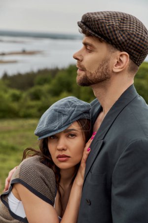 Portrait of trendy brunette woman in vest and vintage outfit hugging bearded boyfriend in jacket and newsboy cap with rural landscape at background, fashionable couple in countryside