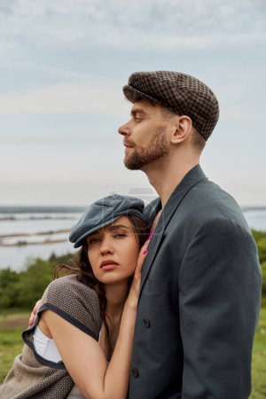 Trendy and brunette woman in newsboy cap and vest hugging bearded boyfriend in jacket and looking at camera with rural landscape at background, fashionable couple in countryside