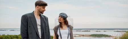 Fashionable brunette woman in vintage outfit looking at bearded boyfriend in jacket and newsboy cap while standing with nature at background, stylish pair amidst nature, banner 