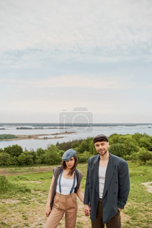 Smiling bearded man in vintage-inspired outfit holding hand of brunette girlfriend in suspenders and newsboy cap and standing with nature at background, stylish pair amidst nature