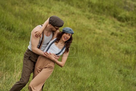 Modischer Mann im Vintage-Outfit und Sonnenbrille umarmt fröhliche Freundin in Hosenträgern und Nachrichtenmütze, während er Zeit miteinander verbringt und auf dem Rasen steht, stylisches Paar inmitten der Natur