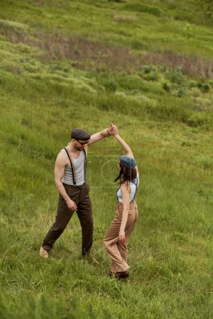 Photo for Smiling bearded man in sunglasses and suspenders having fun with brunette girlfriend in newsboy cap and standing with blurred grassy meadow at background, stylish pair amidst nature - Royalty Free Image
