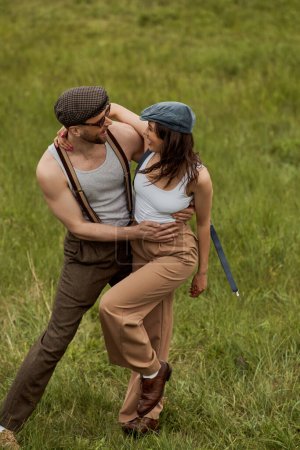 Homme barbu positif et à la mode dans des lunettes de soleil étreignant petite amie brune en bonnet de newsboy et bretelles tout en se tenant debout sur un terrain herbeux à l'arrière-plan, paire élégante au milieu de la nature