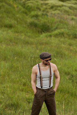 Trendiger Mann im Vintage-Outfit mit Sonnenbrille und Hosenträgern, die Hände in Hosentaschen, während er im Hintergrund auf einer verschwommenen Wiese steht und das Landleben genießt