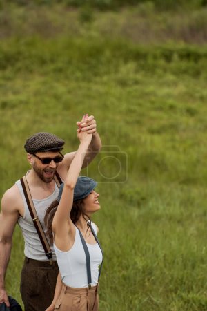 Homme joyeux dans des lunettes de soleil et tenue vintage tenant la main et s'amusant avec une petite amie brune dans une casquette de newsboy et debout sur un champ herbeux flou, couple élégant profitant de la vie à la campagne