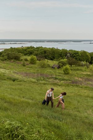 Femme brune en tenue vintage élégante tenant la main du petit ami dans des lunettes de soleil et des bretelles et marchant sur une colline herbeuse avec la nature en arrière-plan, couple élégant profitant de la vie à la campagne