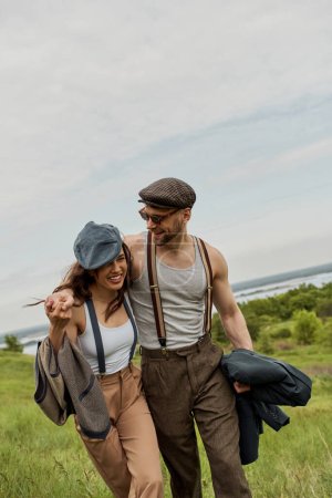 Smiling and bearded man in sunglasses and suspenders hugging brunette girlfriend in vintage outfit and walking together with scenic nature at background, stylish couple enjoying country life