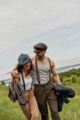 Smiling and bearded man in sunglasses and suspenders hugging brunette girlfriend in vintage outfit and walking together with scenic nature at background, stylish couple enjoying country life Tank Top #663008816