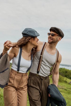 Lächelnder bärtiger Mann im Vintage-Outfit und Kioskmütze umarmt brünette Freundin mit Hosenträgern und spaziert vor malerischer Landschaft im Hintergrund, stilvolles Paar genießt das Landleben