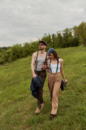 Fashionable man in sunglasses and newsboy cap holding hand of girlfriend in vintage outfit and suspenders and walking on grassy hill and landscape at background, trendy couple in the rustic outdoors