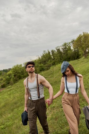 Fashionable bearded an in sunglasses and suspenders holding jacket and hand of brunette girlfriend in newsboy cap and walking on grassy lawn, trendy couple in the rustic outdoors