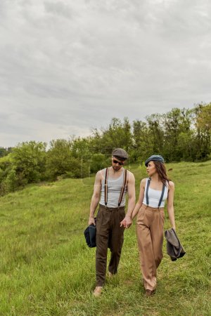 Fashionable brunette woman in newsboy cap and suspenders holding hand of bearded boyfriend in sunglasses and talking while walking together on grassy lawn, trendy couple in the rustic outdoors