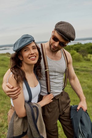 Photo for Fashionable man in sunglasses and suspenders hugging cheerful brunette girlfriend and holding jacket while walking on blurred grassy field, trendy couple in the rustic outdoors - Royalty Free Image