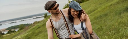 Smiling and fashionable man in sunglasses and suspenders hugging brunette girlfriend in newsboy cap and walking together on grassy field at background, trendy couple in the rustic outdoors, banner 