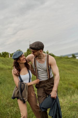 Fashionable bearded man in sunglasses and newsboy cap hugging cheerful brunette girlfriend and holding jacket while walking together in rural landscape, stylish partners in rural escape