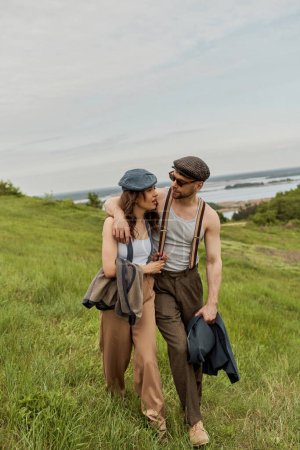 Modischer Mann im Vintage-Outfit und Sonnenbrille umarmt brünette Freundin und spricht beim Spazierengehen auf verschwommenem Grashügel mit bewölktem Himmel im Hintergrund, stilvolle Partner in ländlicher Flucht