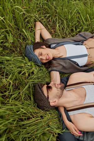 Top view of smiling brunette woman in vintage outfit hugging bearded boyfriend in sunglasses and newsboy cap while lying together on grassy lawn, stylish partners in rural escape