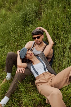 High angle view of fashionable romantic couple in vintage outfits, newsboy caps and suspenders looking at camera while sitting and relaxing on green field, fashionable couple surrounded by nature