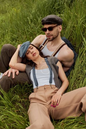 Hombre de moda en gorra de reportero y gafas de sol sentado cerca de novia morena en tirantes y traje vintage en hierba verde y prado, pareja de moda rodeada de naturaleza, escapada romántica