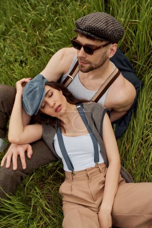 Draufsicht auf modische brünette Frau in Nachrichtenmütze und Vintage-Outfit entspannen in der Nähe bärtigen Freund mit Sonnenbrille auf der Wiese, modische Paar von der Natur umgeben, romantisches Wochenende