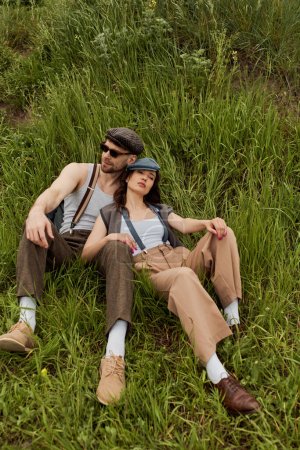 Trendiges romantisches Paar in Vintage-Outfits und Kioskmützen entspannt nebeneinander auf einem Hügel mit grünem Gras im Sommer, modisches Paar umgeben von Natur, romantisches Wochenende