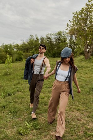 Fashionable brunette woman in vintage outfit and newsboy cap holding hand of bearded boyfriend in sunglasses with jacket and walking on grassy field, fashionable couple surrounded by nature